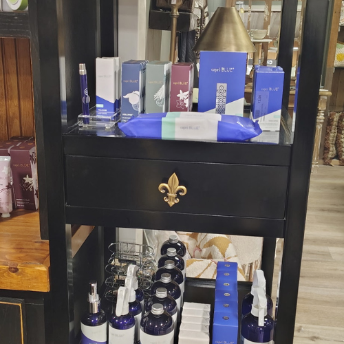 Perfume and beauty products in blue and purple packaging displayed on a black cabinet with a brass fleur-de-lis, creating an elegant boutique feel.