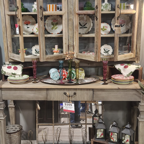 Rustic wooden china cabinet displaying floral dishes, colorful glassware, folded linens, and lanterns in a cozy vintage shop setting.