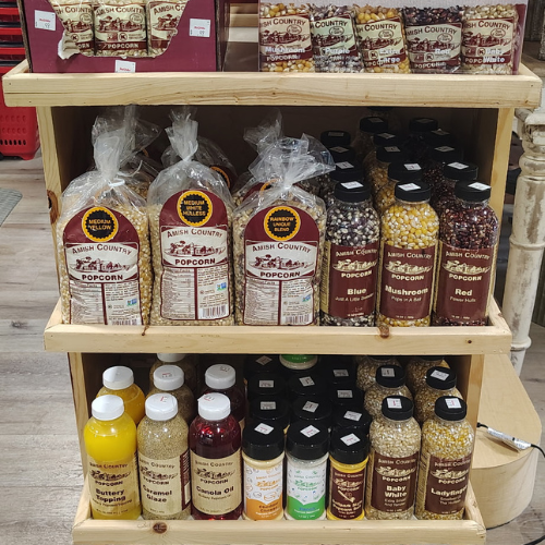 Wooden market shelf displays Amish Country popcorn in colorful bags and jars, including mushroom, blue, red, caramel, and cheddar flavors.