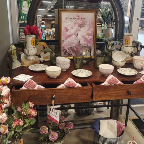 Farmhouse-style console display with floral art, bowls, plates, vases, towels, and pink flowers in a warm, cozy retail setting.