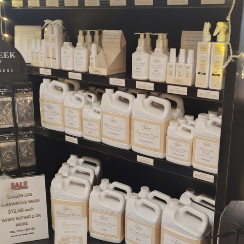 Retail shelf stocked with white gallon jugs and bottles of Glamorous Wash laundry detergent, with a sale sign highlighting bulk savings.