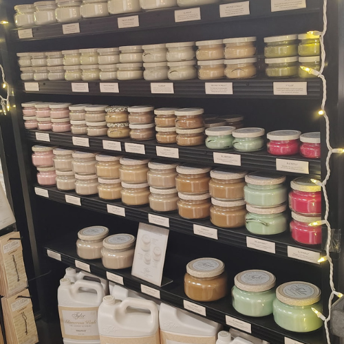 Warmly lit shelves display rows of colorful handmade candles in glass jars and white containers, creating a cozy boutique atmosphere.