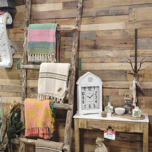 Rustic farmhouse display with colorful woven blankets on a ladder, vintage clock, plants, and pottery arranged against a wood wall.