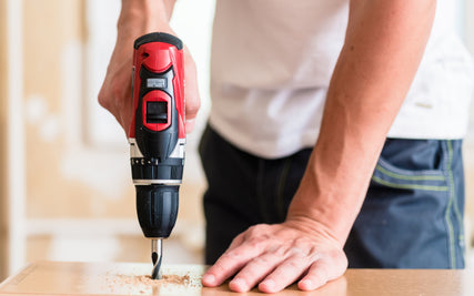 DIY RentalsA person using a red cordless drill to make a hole in a wooden surface, with sawdust scattered. The image conveys focus and precision in DIY work.
