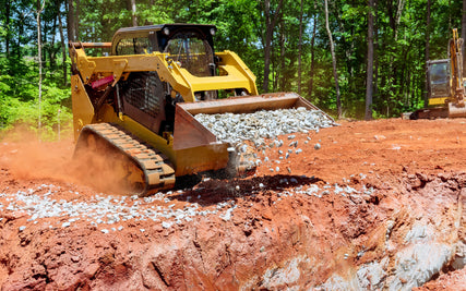 Contractor RentalsA yellow bulldozer spreads gravel over a red dirt construction site, surrounded by green trees. The scene conveys industrial activity in a natural setting.