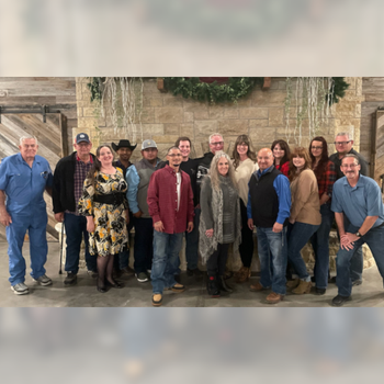 A group of 17 people posing indoors in front of a rustic stone wall with holiday greenery. They appear relaxed and cheerful, dressed in casual attire.
